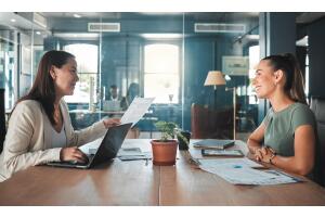 Mulher jovem sendo entrevistada em ambiente corporativo moderno, sentada à mesa com uma recrutadora que analisa seu currículo. A cena transmite profissionalismo, preparação e confiança — ideal para ilustrar conteúdos sobre como fazer um bom currículo.