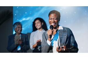 Mulher negra sorrindo enquanto fala ao microfone em uma cerimônia de premiação, segurando um troféu. Ao fundo, duas colegas aplaudem com entusiasmo. A imagem transmite superação, autoconfiança e destaque profissional.