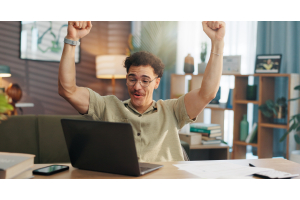Homem jovem de óculos e bigode comemorando em frente a um computador, ele parece estar em home office.