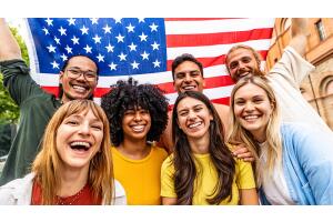 Grupo de jovens sorridentes de diferentes origens, reunidos em frente a uma bandeira dos Estados Unidos, representando diversidade e conexão com o idioma inglês. A imagem reforça a importância do inglês para o mercado de trabalho, especialmente em context