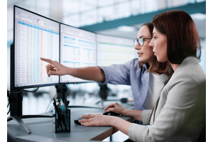 A imagem mostra duas profissionais analisando planilhas em monitores no escritório, representando o ambiente de trabalho típico ao discutir quanto ganha um programador